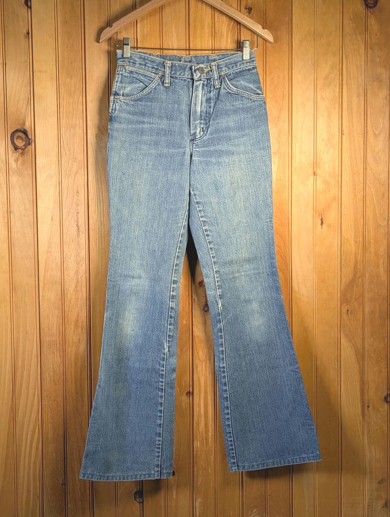 May include: Light blue denim jeans with a flared leg design, hanging on a wooden hanger. The jeans have a classic five-pocket design and a button and zipper closure. The denim has a faded appearance.