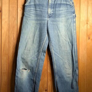 May include: Light blue denim work pants with a distressed knee, hanging on a wooden hanger. The jeans have a button closure, front pockets, and cuffed hems. The background is a wooden wall.