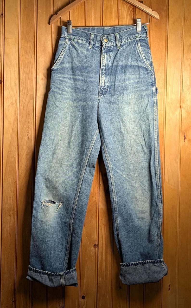 May include: Light blue denim work pants with a distressed knee, hanging on a wooden hanger. The jeans have a button closure, front pockets, and cuffed hems. The background is a wooden wall.
