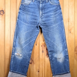 May include: Blue denim jeans with distressed details, including a tear on the left knee. The jeans have a classic five-pocket design and are cuffed at the ankles. They hang on a wooden hanger against a wood-paneled background.