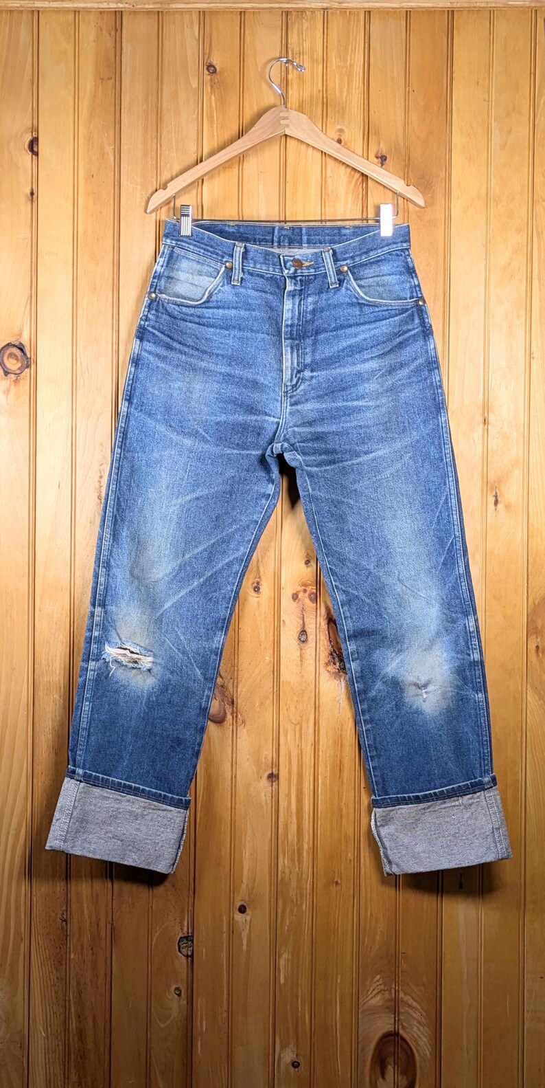 May include: Blue denim jeans with distressed details, including a tear on the left knee. The jeans have a classic five-pocket design and are cuffed at the ankles. They hang on a wooden hanger against a wood-paneled background.