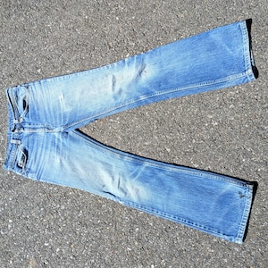 May include: Light blue denim jeans with a faded wash and distressed details. The jeans have a classic five-pocket design, a button and zipper closure, and a straight leg fit. The jeans are laid out on a textured gray surface.