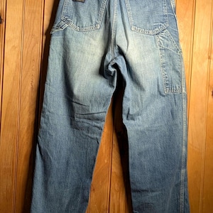 May include: Light blue denim work pants with a faded wash. The pants have a classic five-pocket design, a carpenter pocket, and a small brand patch on the back pocket. The cuffs are rolled up. The pants are hanging on a wooden hanger.