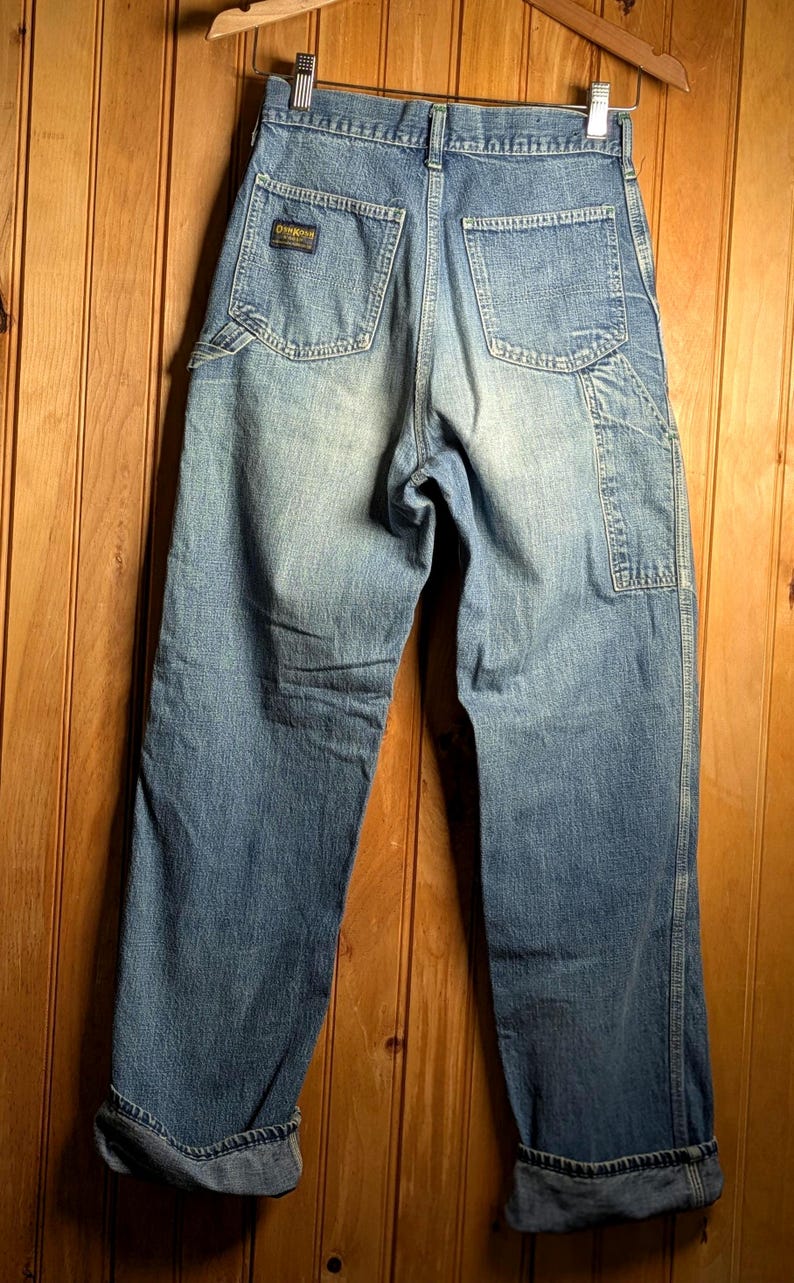 May include: Light blue denim work pants with a faded wash. The pants have a classic five-pocket design, a carpenter pocket, and a small brand patch on the back pocket. The cuffs are rolled up. The pants are hanging on a wooden hanger.