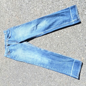 Pode incluir: Um par de calças de ganga azul com lavagem média, corte a direito, design clássico de cinco bolsos, fecho de botão metálico e um detalhe de costura horizontal visível perto da bainha inferior de cada perna, com textura desbotada.
