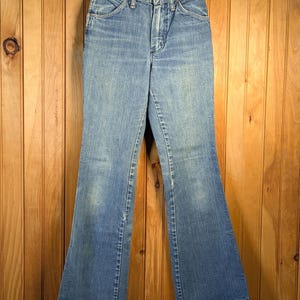 May include: Light blue denim jeans with a flared leg design. The jeans have a classic five-pocket style, a button and zipper closure, and are hanging on a wooden hanger. The jeans are a vintage style.