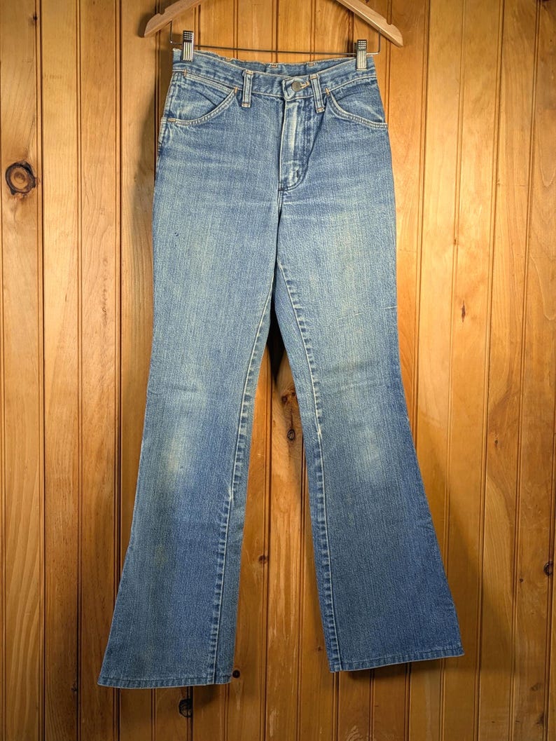 May include: Light blue denim jeans with a flared leg design. The jeans have a classic five-pocket style, a button and zipper closure, and are hanging on a wooden hanger. The jeans are a vintage style.
