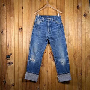 May include: Blue denim jeans with a distressed look, featuring a cuffed hem. The jeans are hanging on a wooden hanger against a wooden panel background. The jeans have a few small holes.
