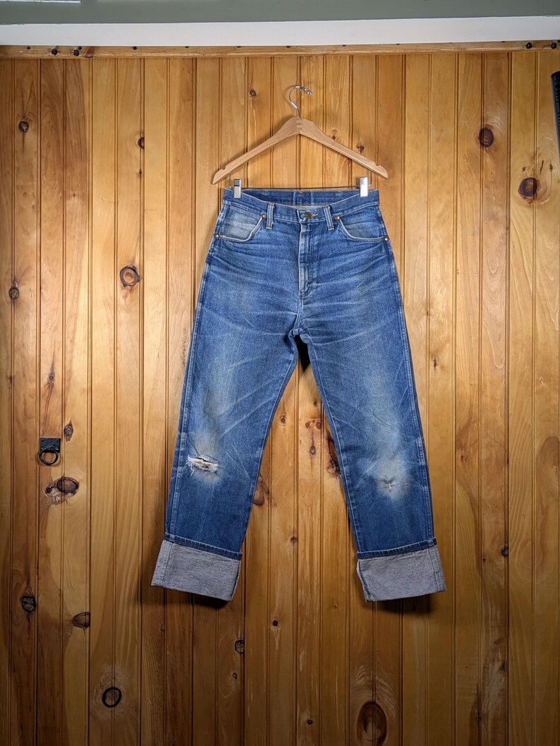 May include: Blue denim jeans with a distressed look, featuring a cuffed hem. The jeans are hanging on a wooden hanger against a wooden panel background. The jeans have a few small holes.