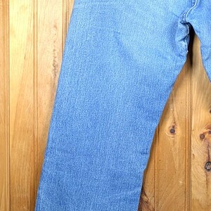 May include: Light blue denim jeans hanging on a wooden hanger. The jeans have a classic five-pocket design with a straight leg. The back pocket features a stitched design. The jeans are made of a durable denim material.
