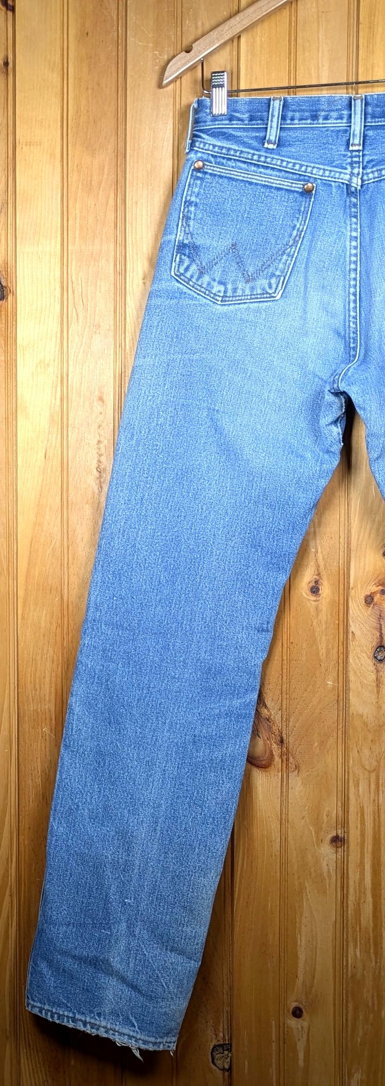 May include: Light blue denim jeans hanging on a wooden hanger. The jeans have a classic five-pocket design with a straight leg. The back pocket features a stitched design. The jeans are made of a durable denim material.