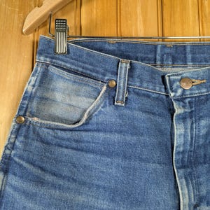 May include: Close-up of blue denim jeans hanging on a wooden hanger. The jeans have a classic five-pocket design with a button and zipper closure. The denim has a faded, worn look.