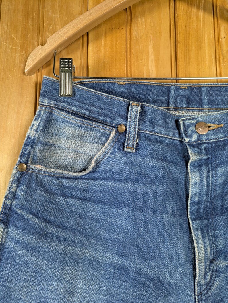 May include: Close-up of blue denim jeans hanging on a wooden hanger. The jeans have a classic five-pocket design with a button and zipper closure. The denim has a faded, worn look.