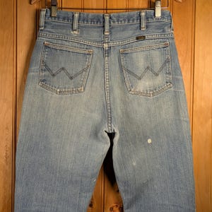 May include: Light blue denim jeans with the Wrangler logo on the back pocket. The jeans are hanging on a wooden hanger against a wooden background. The jeans have a classic five-pocket design.