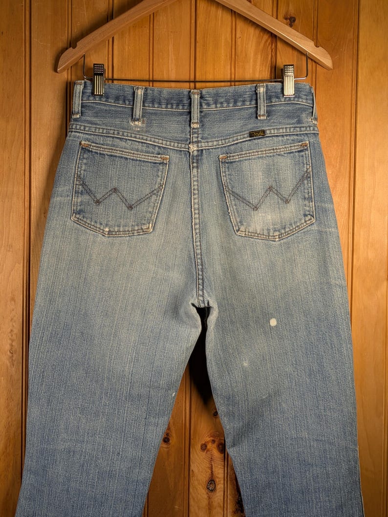 May include: Light blue denim jeans with the Wrangler logo on the back pocket. The jeans are hanging on a wooden hanger against a wooden background. The jeans have a classic five-pocket design.