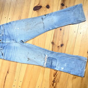 May include: Light blue denim jeans with visible wear and tear, including patches and paint stains. The jeans are laid flat on a wooden surface, showcasing their distressed aesthetic. The jeans have a classic five-pocket design.