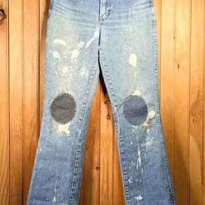 May include: Light blue denim jeans with a high waist and flared legs. The jeans have two dark patches on the knees and white paint splatters. The jeans are hanging on a wooden hanger against a wood-paneled wall.