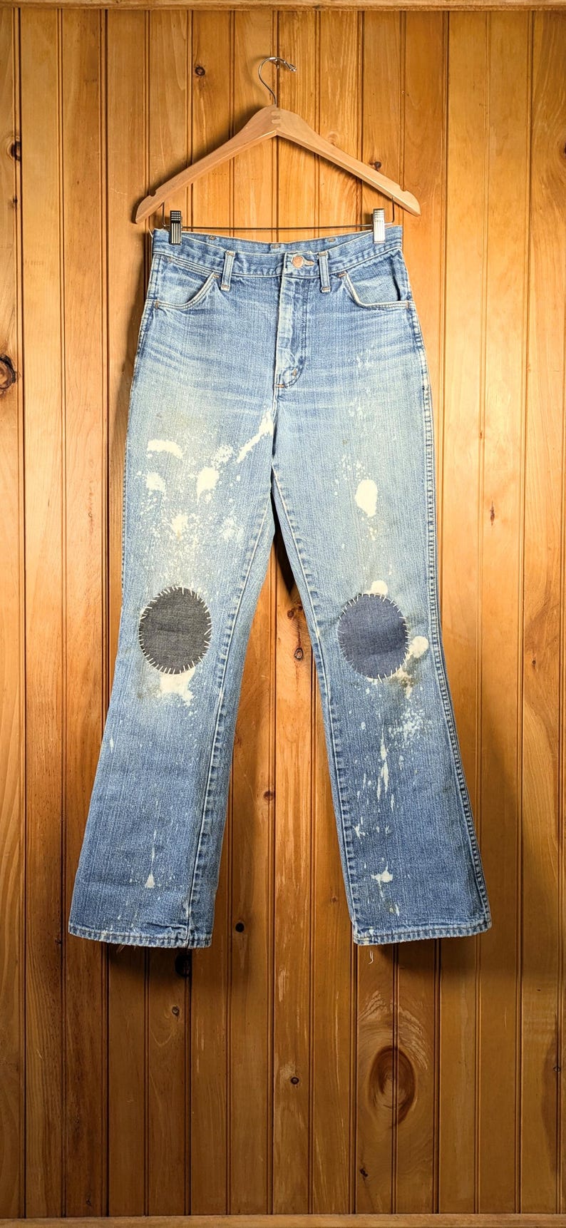 May include: Light blue denim jeans with a high waist and flared legs. The jeans have two dark patches on the knees and white paint splatters. The jeans are hanging on a wooden hanger against a wood-paneled wall.