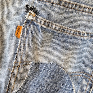 Puede incluir: Primer plano de un bolsillo de vaqueros de mezclilla azul claro. El bolsillo tiene un diseño clásico con una parte inferior curva y costuras reforzadas. Una etiqueta naranja es visible en el bolsillo. Los vaqueros muestran signos de desgaste.