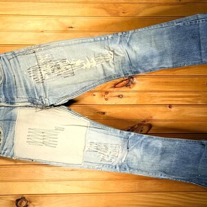 May include: Light blue denim jeans with distressed details and patches, hanging on a wooden hanger. The jeans are laid out on a wooden surface, showcasing their faded wash and unique design elements.