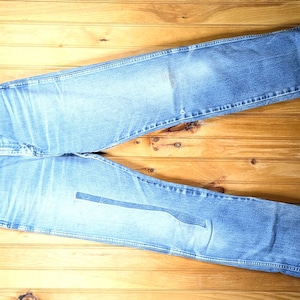 May include: Light blue denim jeans with a classic five-pocket design, laid flat on a wooden surface. The jeans are distressed, showing wear and tear. A wooden hanger is attached to the jeans.