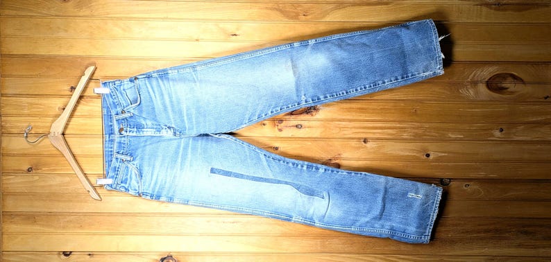 May include: Light blue denim jeans with a classic five-pocket design, laid flat on a wooden surface. The jeans are distressed, showing wear and tear. A wooden hanger is attached to the jeans.