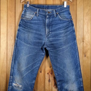 May include: Blue denim jeans with a distressed look, featuring a worn appearance and a ripped knee. The jeans are hanging on a wooden hanger against a wooden background. The jeans have a classic five-pocket design.