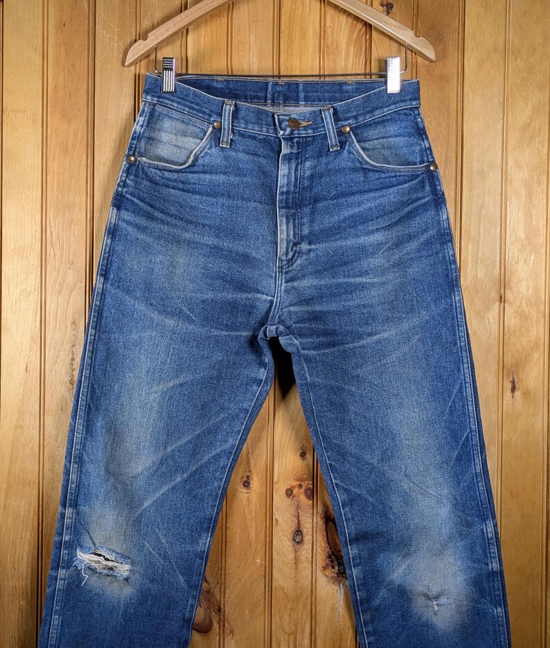 May include: Blue denim jeans with a distressed look, featuring a worn appearance and a ripped knee. The jeans are hanging on a wooden hanger against a wooden background. The jeans have a classic five-pocket design.