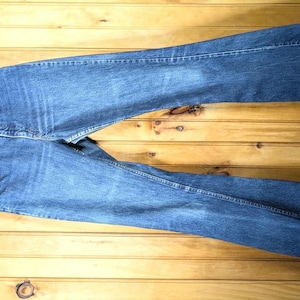 May include: A pair of blue denim jeans hanging on a wooden hanger. The jeans are a medium wash with visible seams and pockets. The background is a wooden surface.
