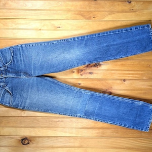 May include: A pair of blue denim jeans hanging on a wooden hanger. The jeans are folded in half and have a light wash.