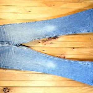 May include: A pair of light blue denim jeans with a wide leg and a flared bottom. The jeans are hanging on a wooden hanger against a wood panel background.