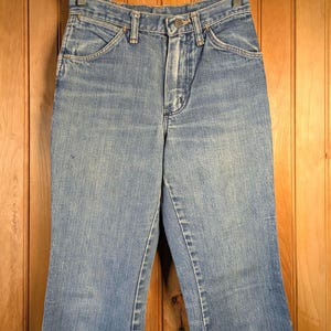 May include: Light blue denim jeans with a classic five-pocket design. The jeans have a high waist and a slight flare at the hem. They are hanging on a wooden hanger against a wooden background.