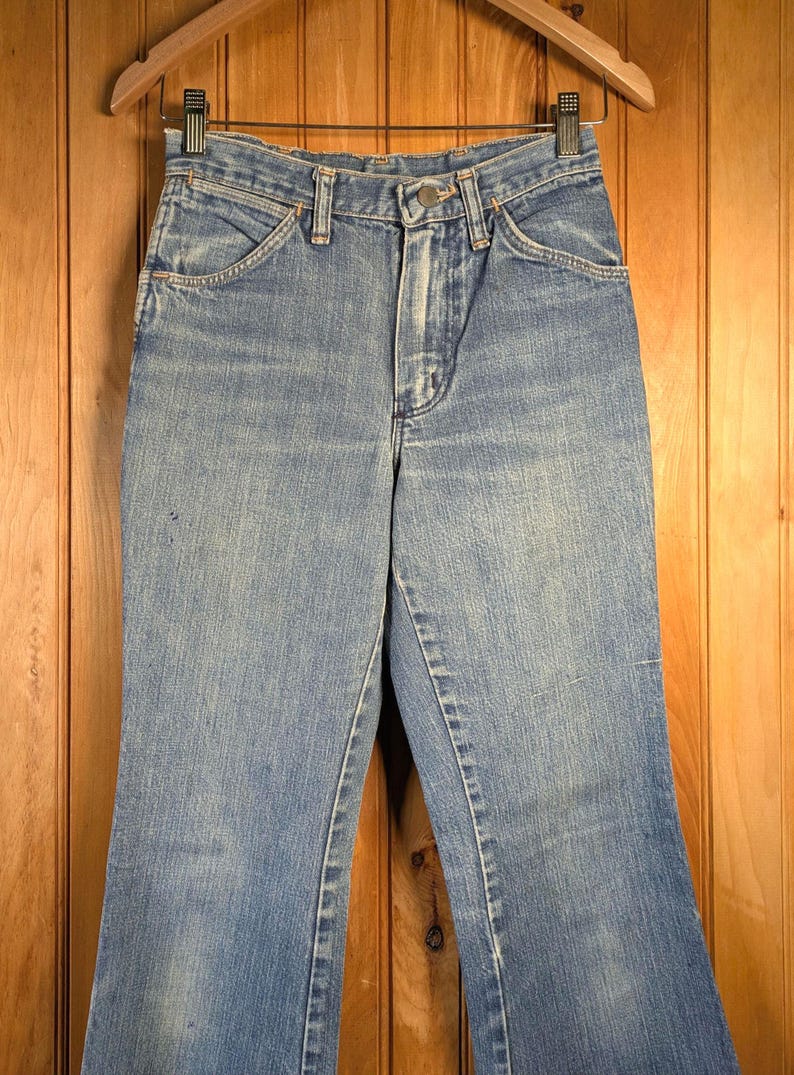 May include: Light blue denim jeans with a classic five-pocket design. The jeans have a high waist and a slight flare at the hem. They are hanging on a wooden hanger against a wooden background.