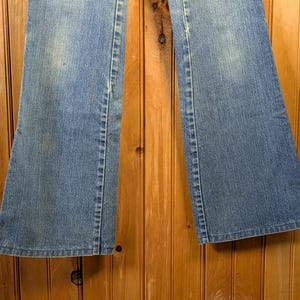 May include: Light blue denim jeans with a flared leg design. The jeans have a classic five-pocket style and a faded wash. The seams are visible, and the fabric appears to be slightly worn. The jeans are displayed against a wooden background.