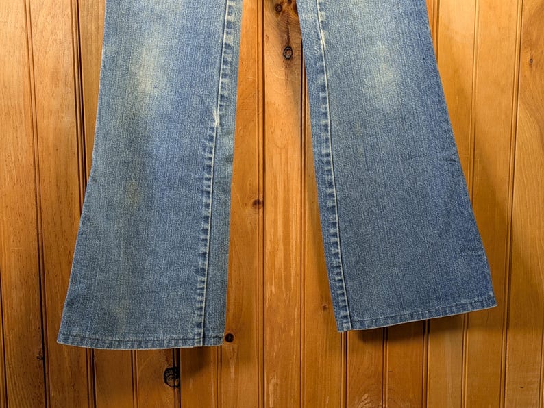 May include: Light blue denim jeans with a flared leg design. The jeans have a classic five-pocket style and a faded wash. The seams are visible, and the fabric appears to be slightly worn. The jeans are displayed against a wooden background.