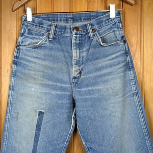 May include: Light blue denim jeans with a faded appearance, featuring a patch on the left leg. The jeans are hanging on a wooden hanger against a wooden background. The jeans have a button and zipper closure.