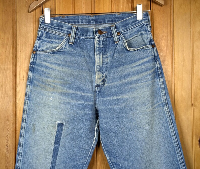 May include: Light blue denim jeans with a faded appearance, featuring a patch on the left leg. The jeans are hanging on a wooden hanger against a wooden background. The jeans have a button and zipper closure.
