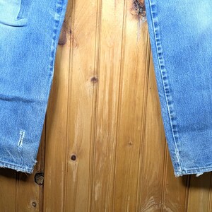 May include: Light blue denim jeans with distressed details, including a tear near the hem. The jeans have a classic five-pocket design and a faded appearance. The bottom hems are frayed.