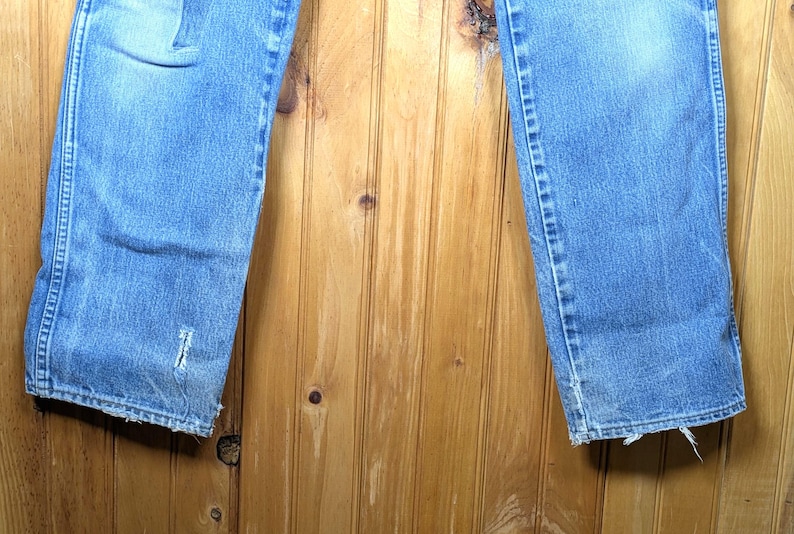 May include: Light blue denim jeans with distressed details, including a tear near the hem. The jeans have a classic five-pocket design and a faded appearance. The bottom hems are frayed.