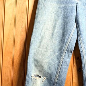 May include: Light blue denim jeans with a distressed knee and cuffed hem. The jeans are hanging on a wooden hanger against a wood-paneled wall. The jeans have a classic five-pocket design.