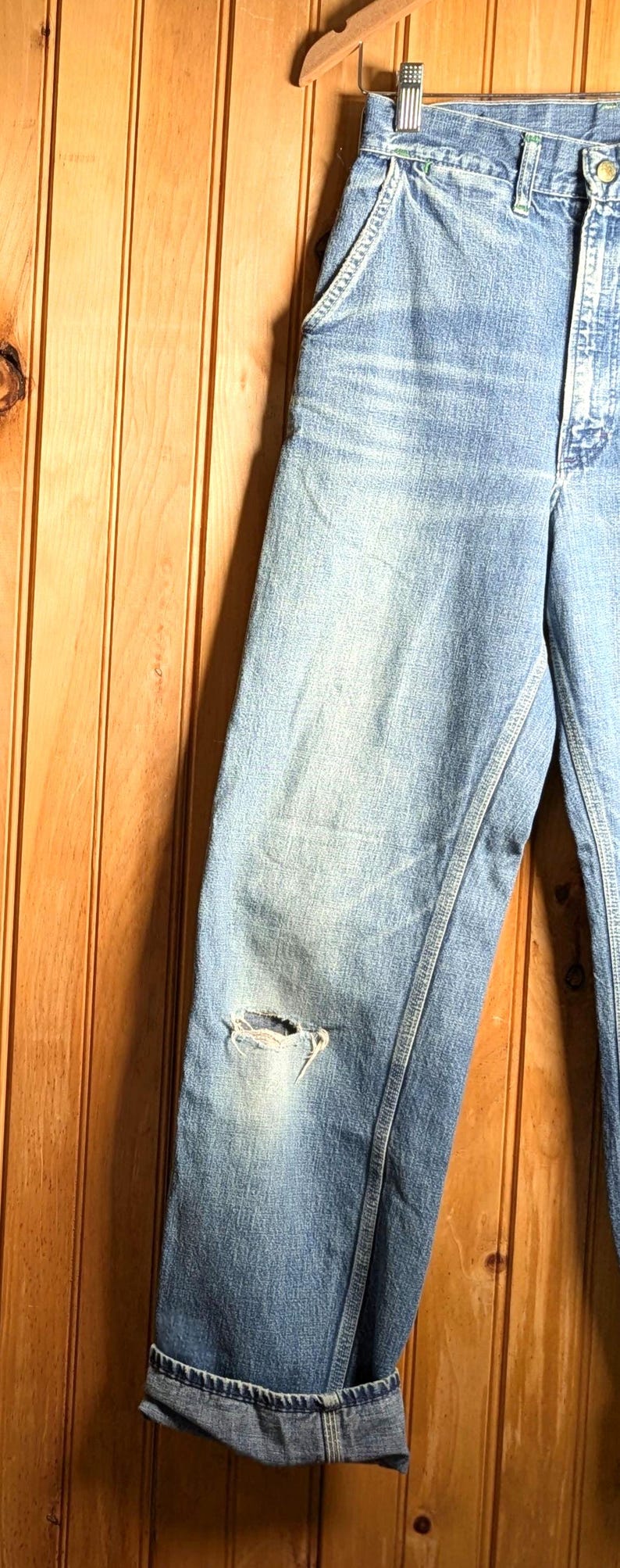 May include: Light blue denim jeans with a distressed knee and cuffed hem. The jeans are hanging on a wooden hanger against a wood-paneled wall. The jeans have a classic five-pocket design.