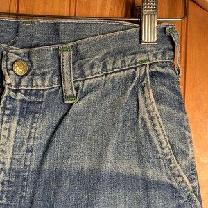 May include: Close-up of blue denim jeans with green stitching accents. The jeans feature a button closure and a pocket with green trim. The garment is hanging on a metal hanger against a wooden background.