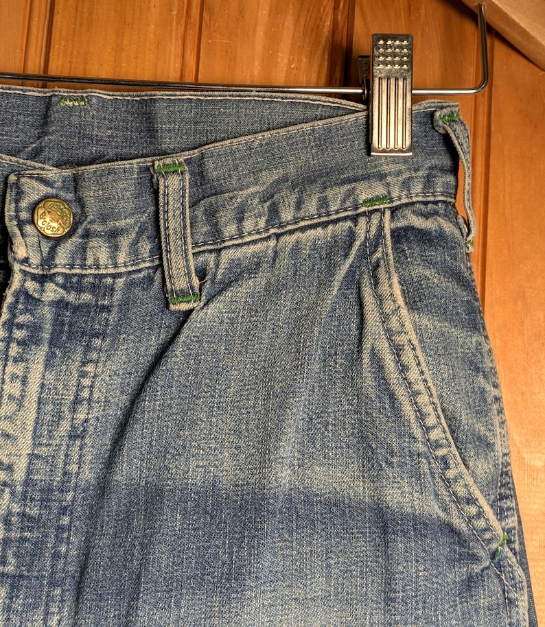 May include: Close-up of blue denim jeans with green stitching accents. The jeans feature a button closure and a pocket with green trim. The garment is hanging on a metal hanger against a wooden background.