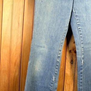 May include: Light blue denim jeans hanging on a wooden hanger. The jeans have a classic five-pocket design and a slight flare at the bottom. The denim has some fading and wear, giving them a vintage look.