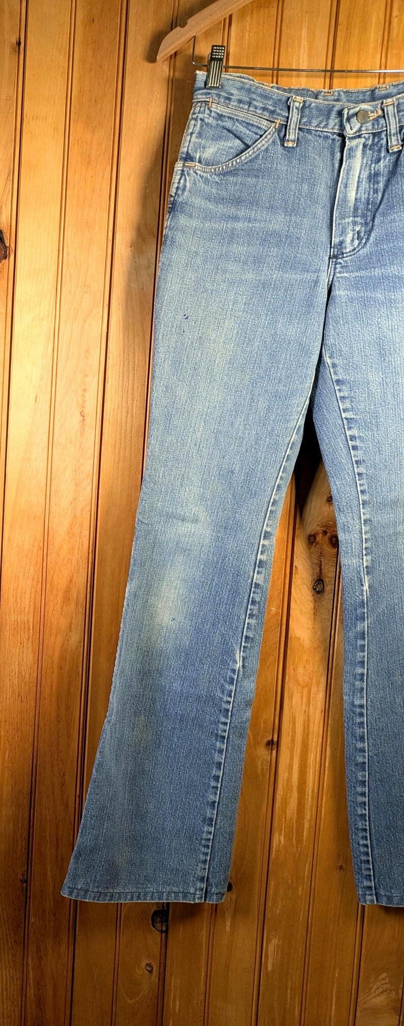 May include: Light blue denim jeans hanging on a wooden hanger. The jeans have a classic five-pocket design and a slight flare at the bottom. The denim has some fading and wear, giving them a vintage look.