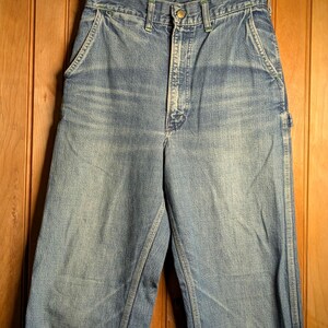 May include: Light blue denim work pants with a distressed look, featuring a worn area on the left knee. The pants have a classic five-pocket design, a button and zipper closure, and are hanging on a wooden hanger.