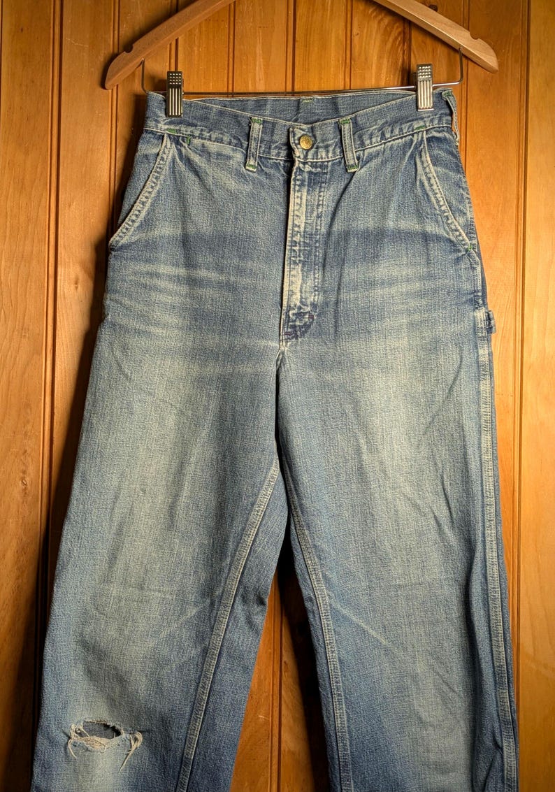 May include: Light blue denim work pants with a distressed look, featuring a worn area on the left knee. The pants have a classic five-pocket design, a button and zipper closure, and are hanging on a wooden hanger.