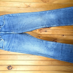 May include: Light blue denim jeans with a classic five-pocket design, hanging on a wooden hanger. The jeans have a faded wash and distressed details, with a straight leg and a relaxed fit. The background is a wooden surface.