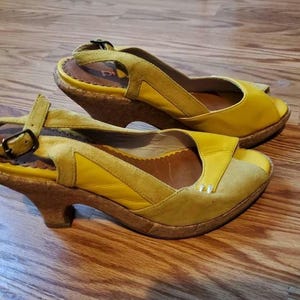 Puede incluir: Un par de sandalias de cuña de cuero amarillo con tacones de corcho. Las sandalias tienen un dedo del pie abierto y una correa de tobillo ajustable.