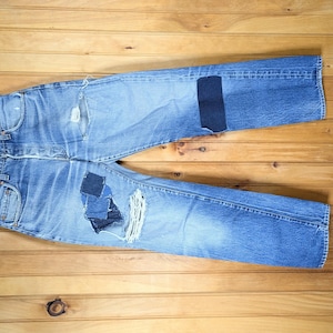 May include: A pair of blue denim jeans with multiple patches sewn on. The jeans are hanging on a wooden hanger against a woodgrain background.
